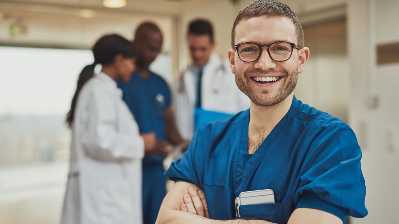 A confident nurse in scrubs smiling, with colleagues talking in the background