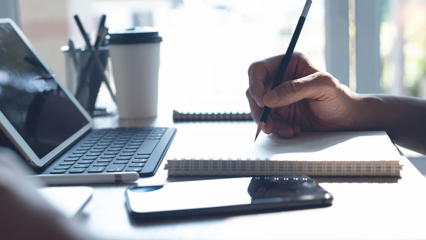 A hand drafting notes in a notebook beside a tablet and keyboard